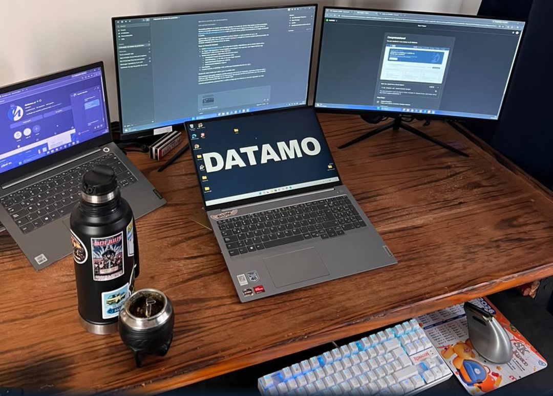El escritorio de DATAMO: tres pantallas, una notebook con el logo y un mate.